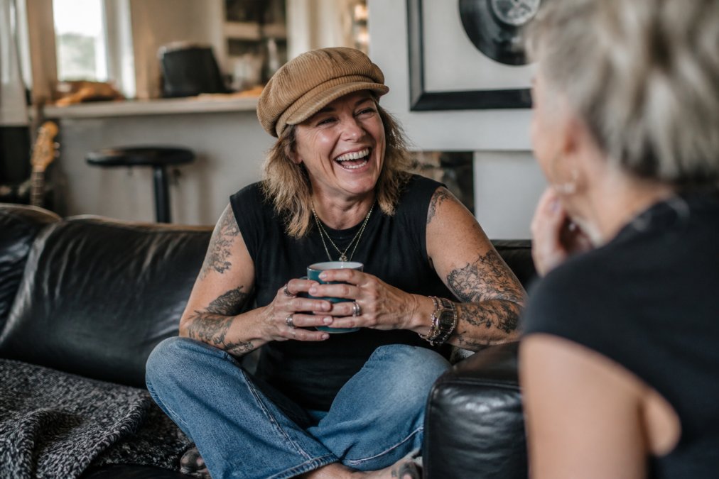 A woman in her late 40s laughing on a couch in casual clothes and a corduroy cap, holding a mug, with a relaxed and rebellious energy, no beige or clinical wellness props in sight