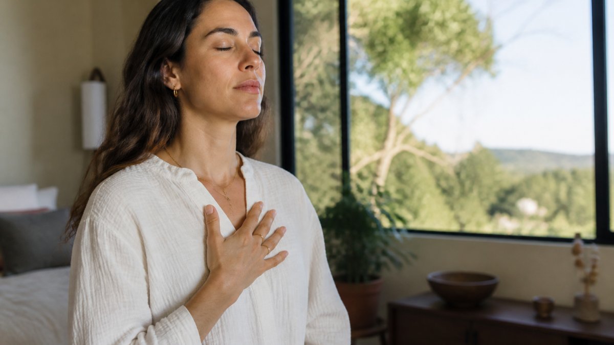 Person sitting in a calm, comfortable space with soft natural lighting, hand placed gently on their chest, eyes closed in a moment of mindful self-awareness and peaceful somatic presence
