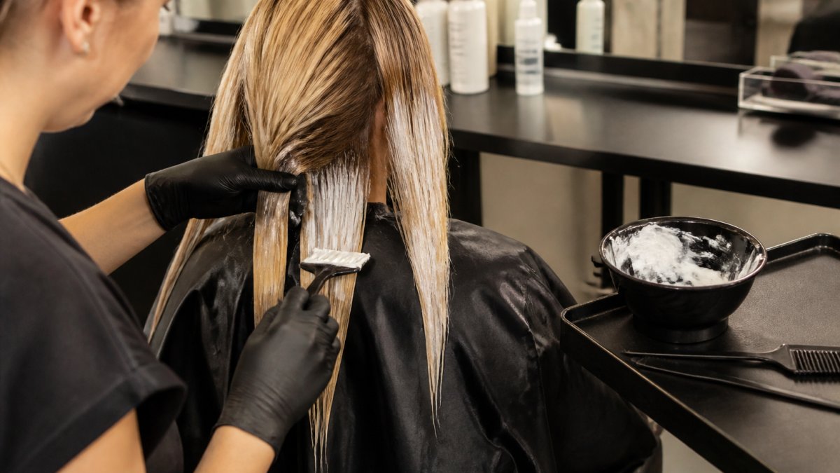 Professional stylist applying a creamy reconstruction mask to bleached blonde hair in a sunlit salon
