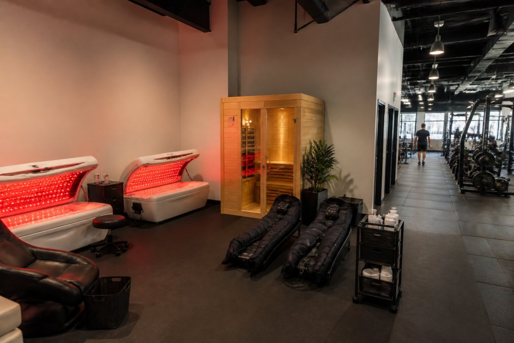 Interior of a small gym with a dedicated recovery zone showing red light therapy beds, a sauna, and compression therapy equipment arranged efficiently, with clear traffic flow between recovery devices and main gym floor, professional lighting, and organized member scheduling