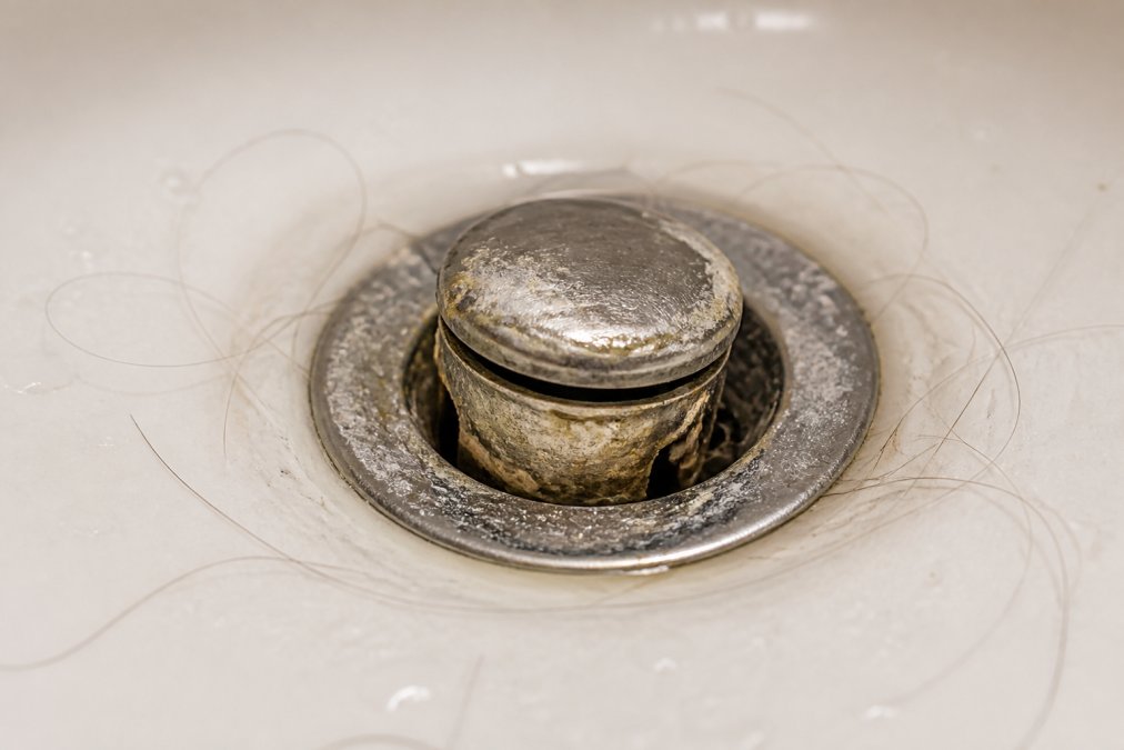 Close-up of a bathroom sink drain with visible hair strands, soap scum buildup, and mineral deposits around the pop-up stopper assembly in natural lighting