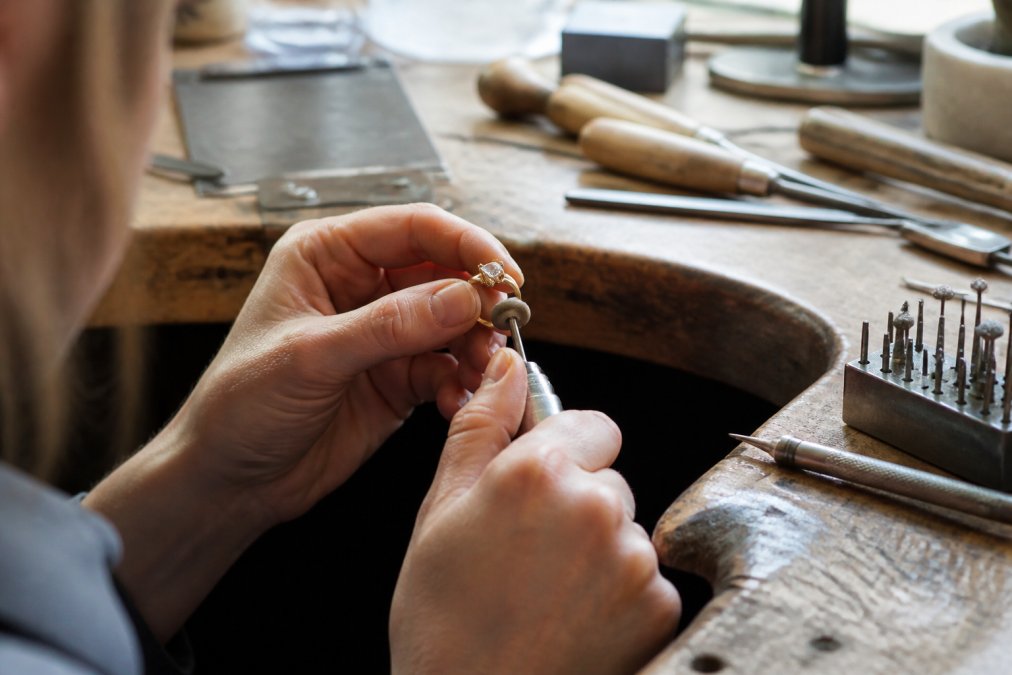 Artisan jeweller's hands polishing and inspecting a handcrafted recycled gold ring on a workbench, with soft natural light highlighting the metal's lustre and fine detail work