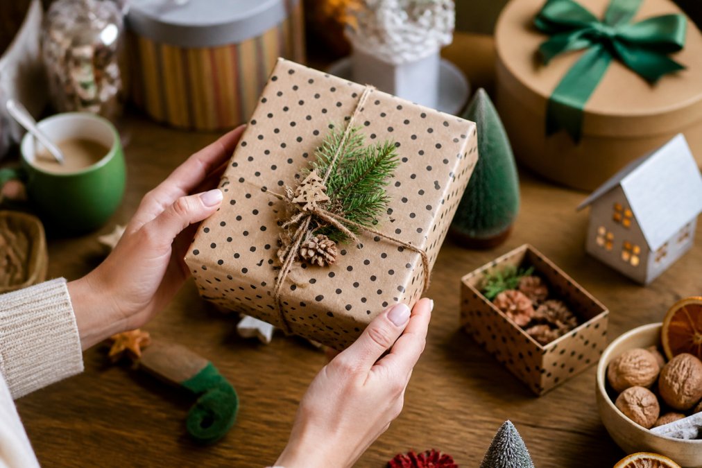 Close-up of hands holding a wrapped gift box with decorative paper and ribbon, with seasonal home decor visible in soft-focus background, suggesting gift-giving and home styling, warm indoor lighting