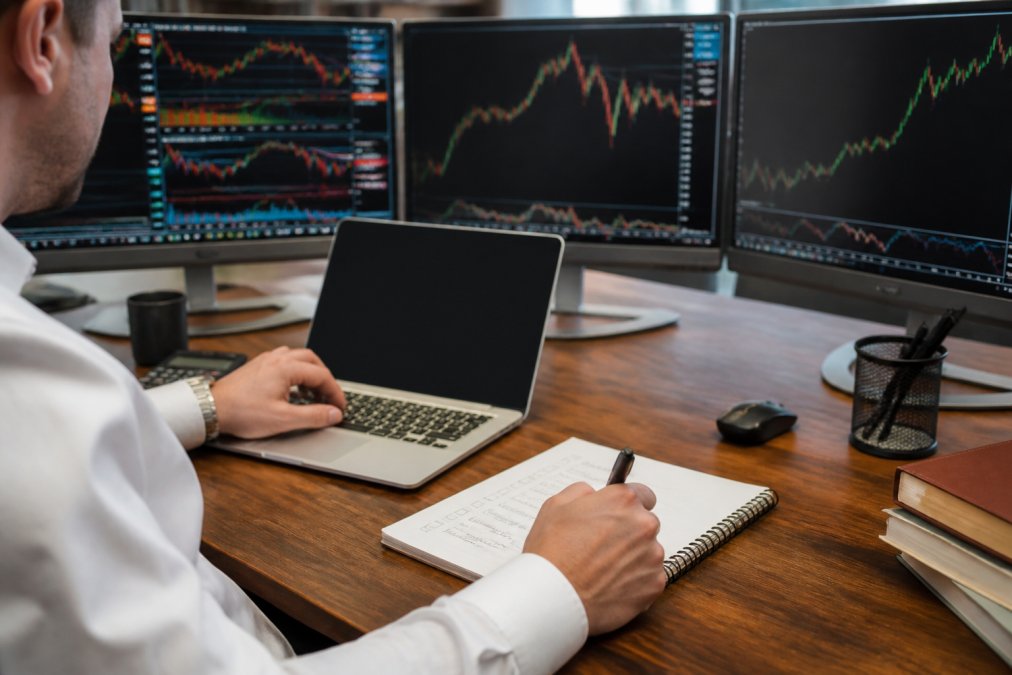Professional trader's hands at laptop with multiple monitors displaying trading charts and software interfaces, with a notepad and checklist visible on desk