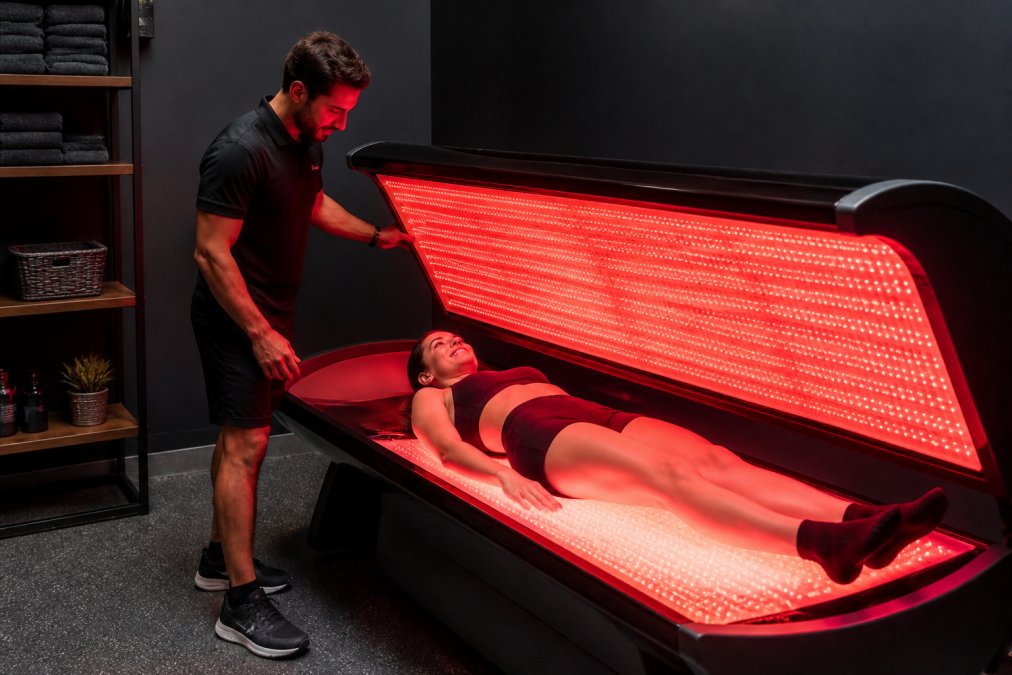 Gym staff member professionally assisting a member during red light therapy session in modern recovery space, showing proper positioning and attentive service delivery in commercial fitness setting