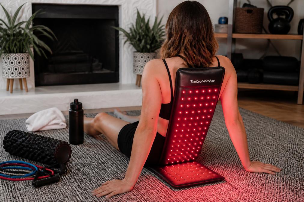 Athlete in home gym setting using red light therapy device on lower back while foam roller and resistance bands are visible on floor, with recovery-focused environment showing water bottle and towel
