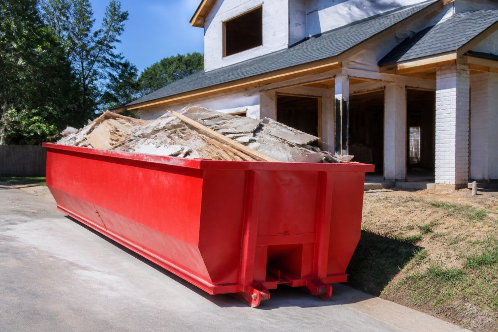 A 15-yard roll-off dumpster parked on a residential driveway filled with construction debris including wood scraps and drywall, a house under renovation visible in the background under clear afternoon light