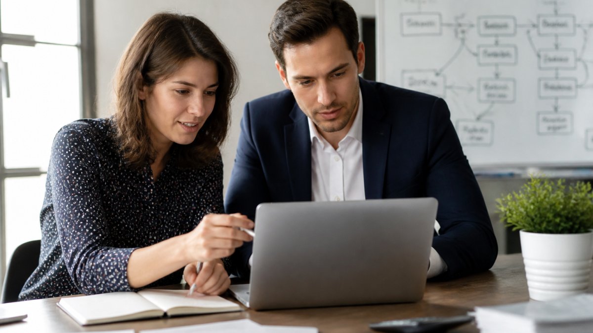 A small business owner and an operations manager reviewing a workflow diagram on a laptop in a bright, modern office, with a whiteboard showing process steps in the background