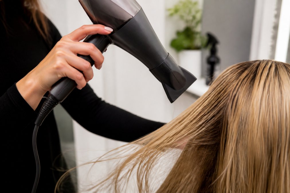 Close-up of hands holding a professional blow dryer with concentrator nozzle attachment pointed downward at a section of damp blonde hair, showing proper 45-degree angle and positioning