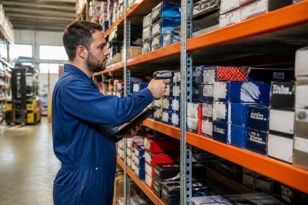 Warehouse worker scanning automotive parts boxes on metal shelving racks in a well-lit auto parts distribution center, tablet in hand