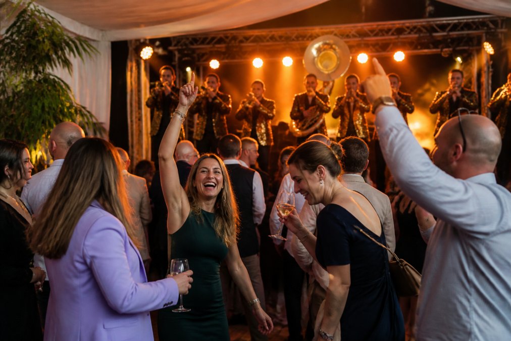 Groep gasten die energiek genieten van live optreden op evenement, dansend en lachend met brassband op podium in feestelijke sfeer