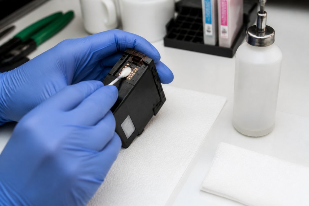 Close-up of hands wearing nitrile gloves using a lint-free swab to clean the copper contact pads and nozzle plate of a refillable ink cartridge on a white workbench, with isopropyl alcohol bottle and paper towels nearby
