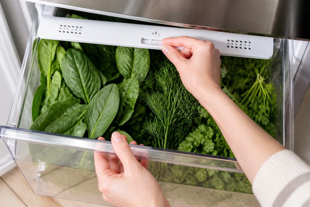 Hands adjusting a humidity slider on a crisper drawer filled with fresh spinach for better refrigerator organization.