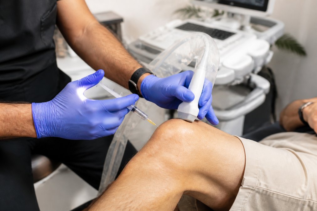 Healthcare provider administering regenerative medicine injection to patient's knee in clinical setting with ultrasound guidance equipment visible, showing minimally invasive procedure technique