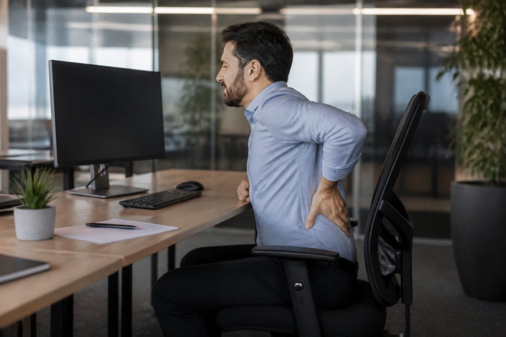 An office worker in his 30s sitting at a desk, rubbing his lower back in discomfort, with a computer monitor and ergonomic chair visible in a modern office setting