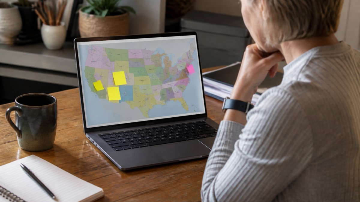 A small business owner reviewing a map of the United States on a laptop screen with sticky notes marking different states, sitting at a wooden desk with a coffee mug and notebook nearby