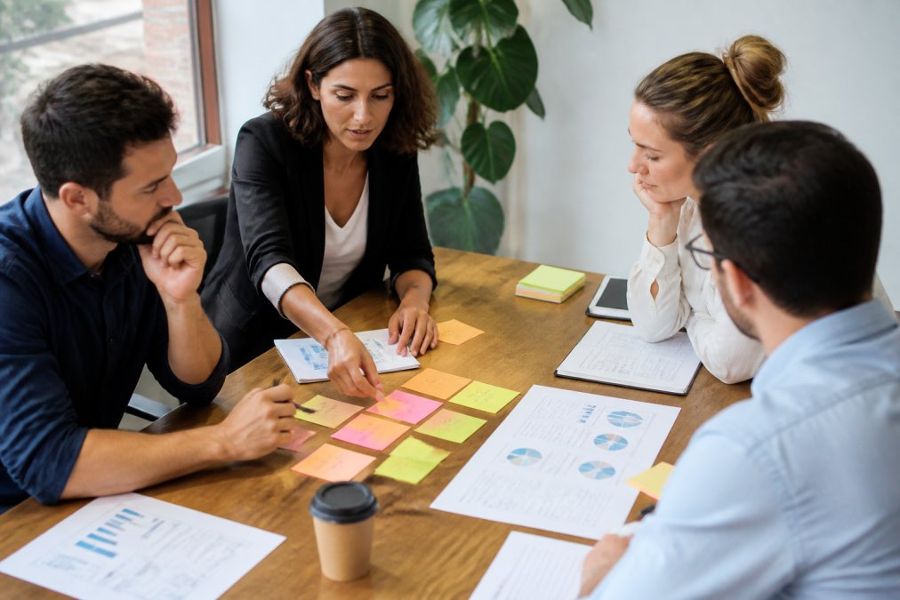 Equipo de marketing colaborando alrededor de un escritorio con notas adhesivas de colores, laptop mostrando un dashboard de analytics y documentos de estrategia visible