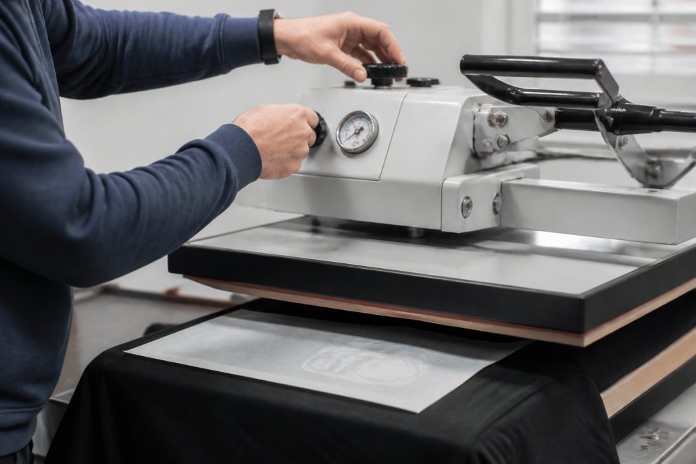 Hands adjusting pressure settings on a heat press control panel, with a pressure gauge visible and transfer film ready for application