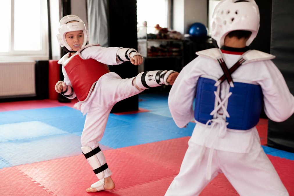 Young martial arts student wearing properly fitted protective gear including headgear, mouthguard, chest protector, and shin guards during supervised training session in bright dojo