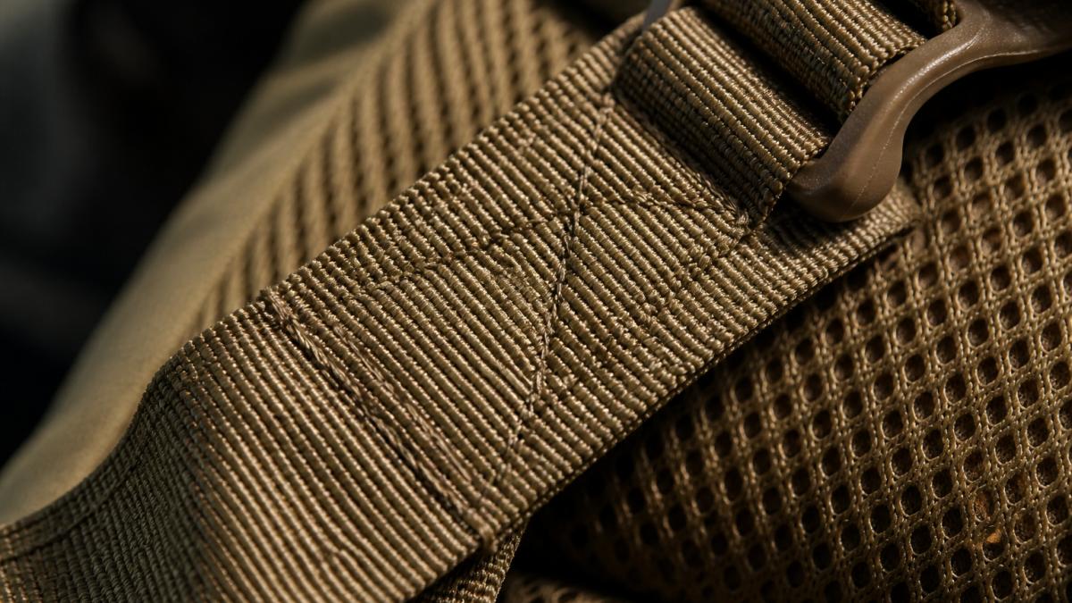 Close-up of reinforced bar tack stitching on a tactical backpack strap showing dense, overlapping stitch pattern under bright natural lighting with visible thread resistance