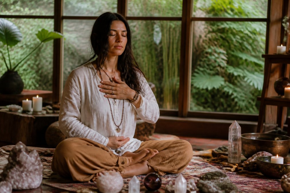 Person sitting cross-legged in peaceful meditation space with soft natural light, surrounded by crystals and calming elements, engaged in mindful reflection