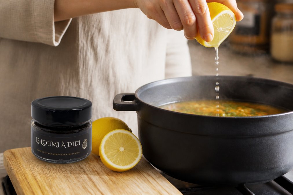 Close-up of hands squeezing fresh lemon juice over a steaming pot of soup, with lemon halves and a glass jar of citron paste on a wooden cutting board, natural kitchen lighting