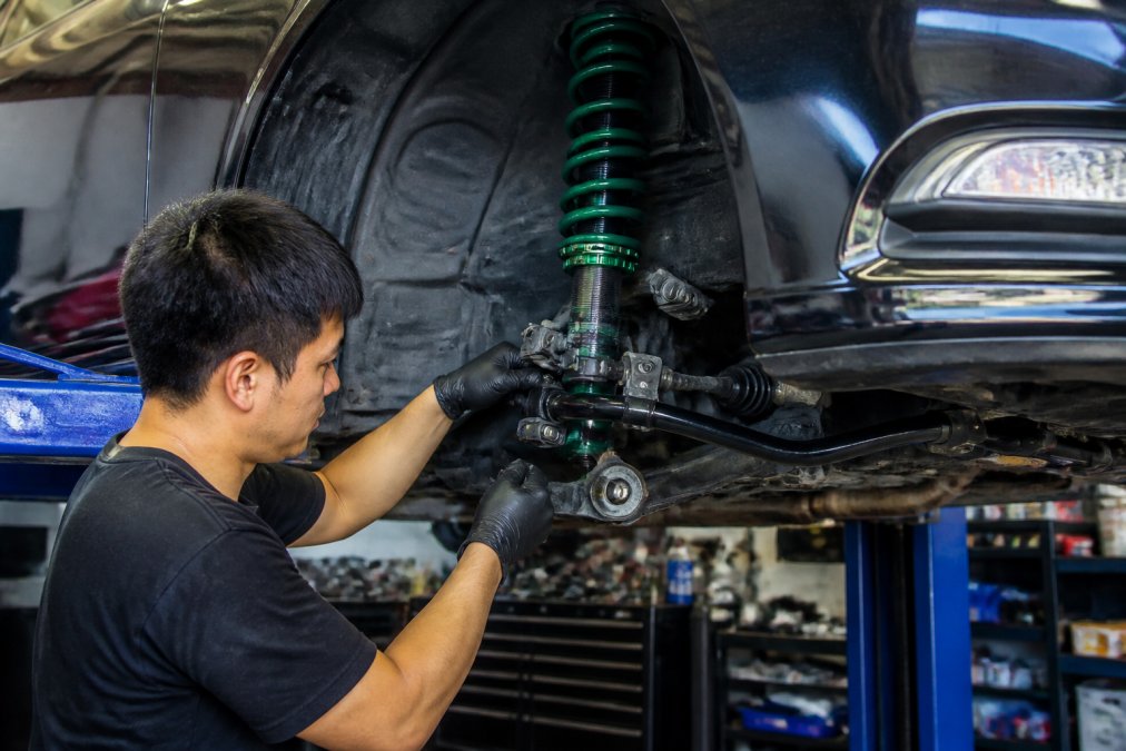 Professional mechanic installing coilover suspension components on a lifted Japanese sports car in workshop bay, showing sway bar and lower control arm installation with precision tools