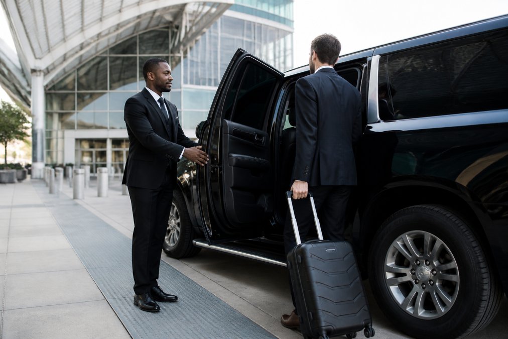 Professional driver in dark suit opening rear door of luxury black truck for business executive with carry-on luggage outside modern airport terminal with glass and steel architecture