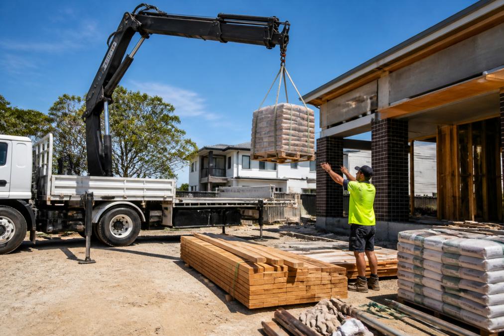 A Hiab crane truck unloading pallets of building materials onto a residential construction site in Sydney, with a worker in a hi-vis vest guiding the crane arm under bright midday sunlight, stacked timber and bagged cement visible in the background