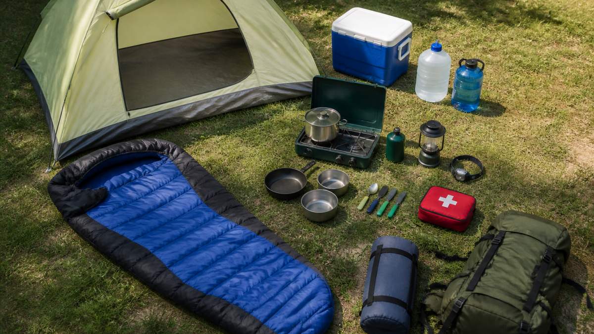 Fully organized campsite viewed from above showing a pitched tent, sleeping bag laid out, camp stove with cookware nearby, blue cooler, water containers, headlamp, and first aid kit arranged on grass with natural daylight