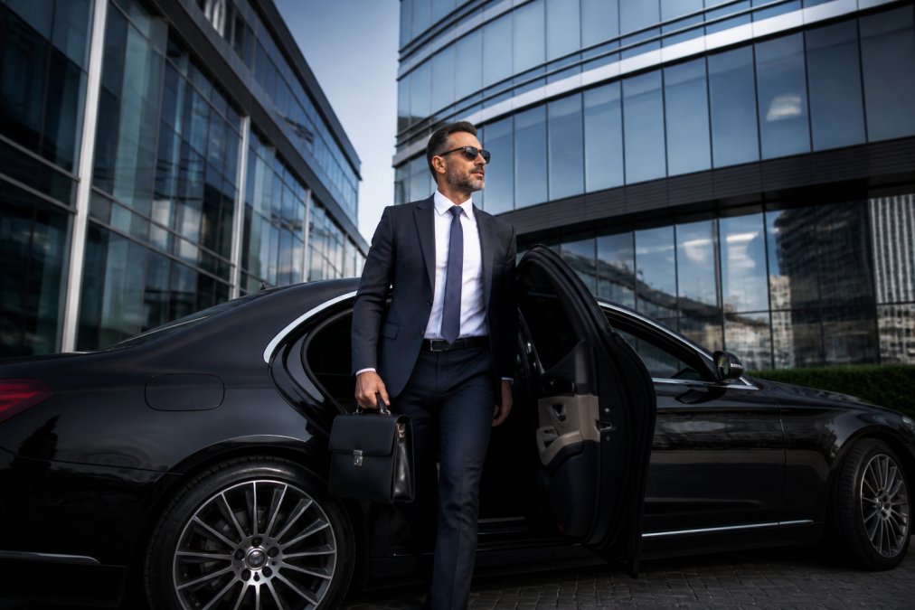Professional business executive in dark suit stepping out of a luxury black sedan in front of a modern glass office tower, briefcase in hand, appearing confident and composed