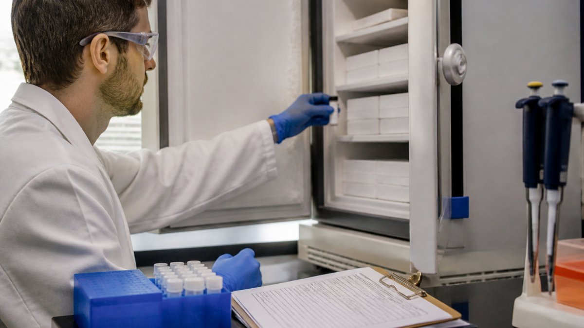 A laboratory researcher in a white coat and gloves placing a labeled peptide vial into a temperature-controlled freezer, with a clipboard and COA documents on the bench nearby