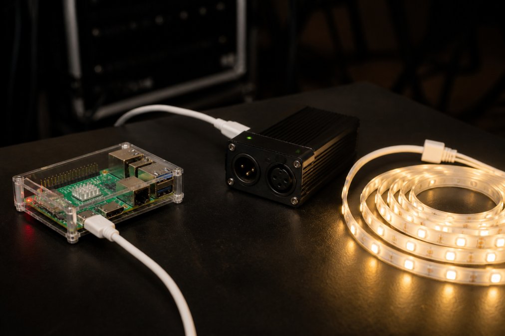 Close-up of a Raspberry Pi 5 connected via USB cable to a compact DMX interface, with a Govee LED strip light coiled on the desk beside it, all lit in warm white against a dark production workspace