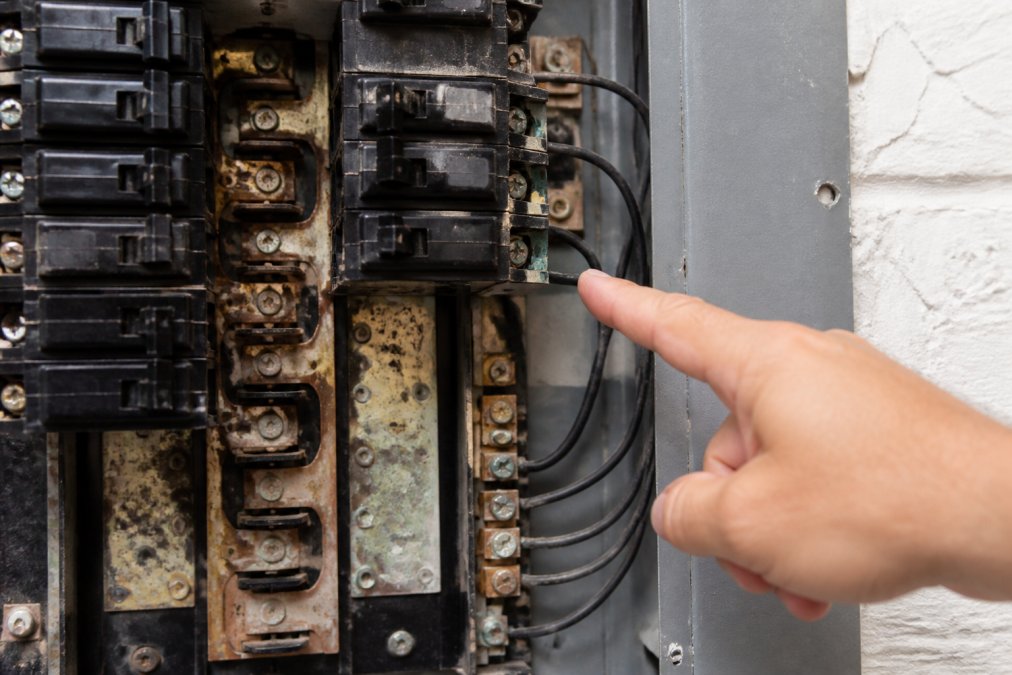 Close-up of an older electrical panel showing worn breakers, corrosion, and discoloration on the bus bars, with a homeowner's hand pointing to areas of visible deterioration