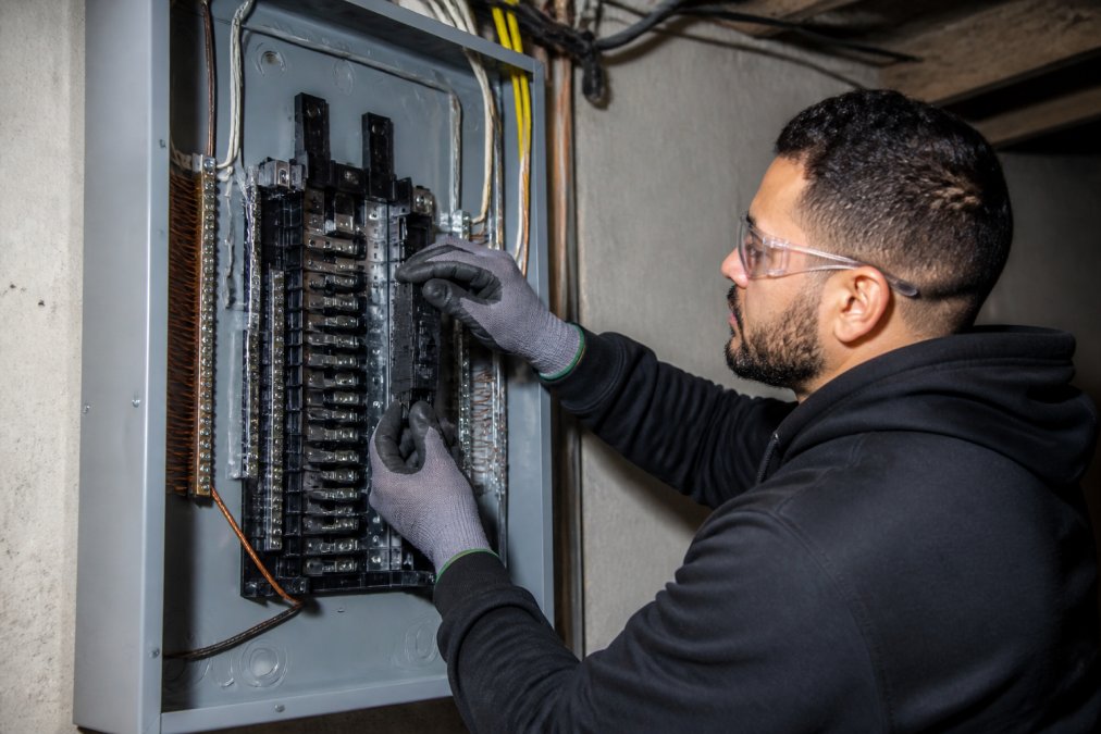 Licensed electrician in safety glasses and work gloves carefully installing circuit breakers into a new electrical panel, with the panel door open showing the bus bars and grounding connections, in a residential basement setting with proper lighting