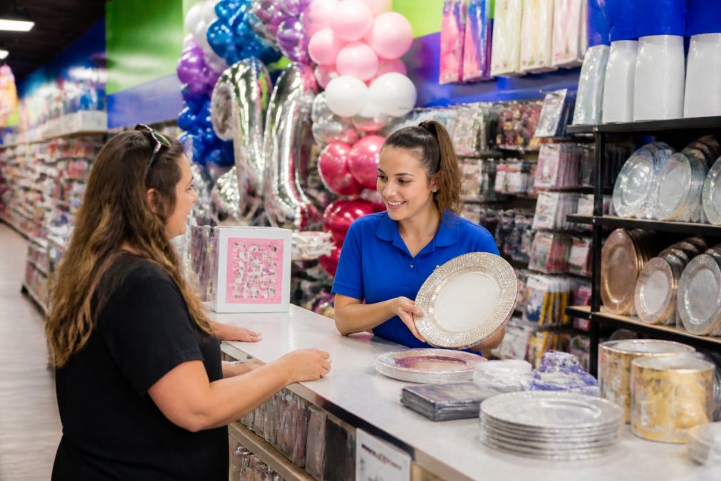 A bright, colorful party supply store interior with rows of balloon arrangements, tableware displays, and rental equipment, a friendly staff member in a branded shirt helping a customer select items at a well-lit service counter