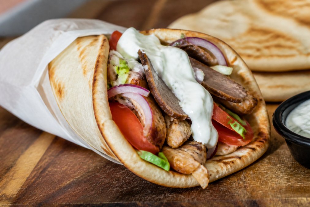Close-up of a freshly made gyro wrap with seasoned chicken, sliced lamb, and crisp vegetables on a wooden serving board, garnished with a generous dollop of tzatziki and warm pita bread under soft natural light