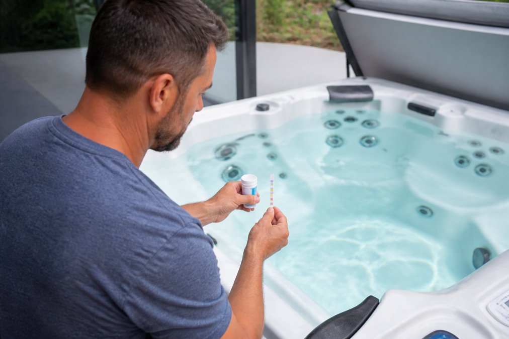 Homeowner in casual clothing checking hot tub water chemistry with test strips, examining the water clarity and color in bright daylight from above the tub