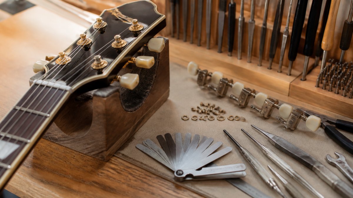 Professional luthier's workbench with organized precision hand tools, tuning machines, feeler gauges, and a guitar headstock positioned for setup work, showing meticulous tool arrangement under warm workshop lighting