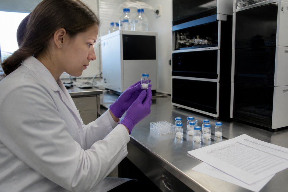 Laboratory technician in white coat examining clear peptide vials under bright analytical lighting, with HPLC equipment and printed batch documentation visible on the stainless steel workbench