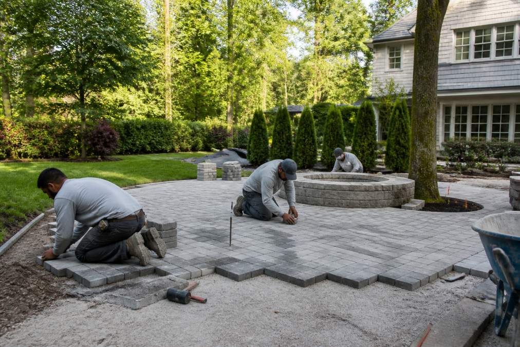 A professional landscaping crew installing a paver patio in a lush, upscale backyard in Westchester County, with mature trees and a manicured lawn