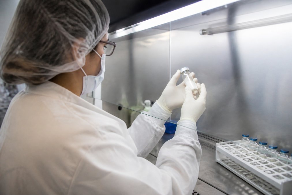 Laboratory technician in sterile gloves handling a vial of lyophilized powder under a biosafety cabinet with temperature monitoring display visible on the wall