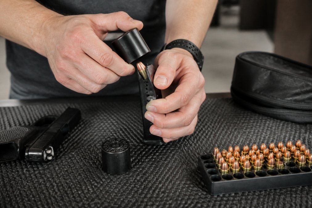 Shooter's hands demonstrating proper grip technique while pressing a 9mm round into a double-stack pistol magazine using a speed loader on a black rubber range mat under indoor range lighting