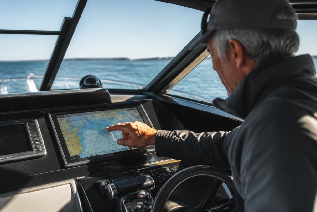A captain's weathered hand tapping a high-resolution touchscreen chartplotter at the helm, calm blue water visible through the windshield ahead