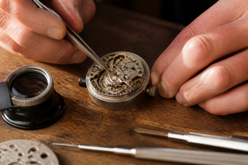 Close-up of a watchmaker's hands using fine tweezers to place a tiny jewel into a mechanical watch movement on a wooden workbench, loupe resting nearby, warm task lighting