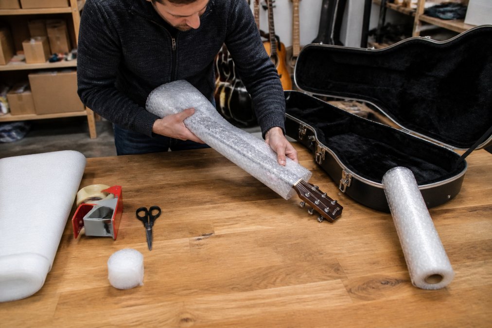 Person carefully wrapping a guitar neck with bubble wrap on a wooden workbench, with a hard-shell case open beside them and packing materials spread around