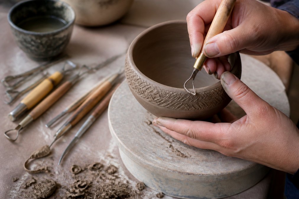 Potter using a V-tip palm carver to etch intricate patterns into a leather-hard clay bowl with professional pottery tools.