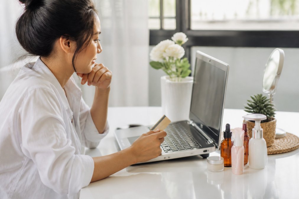 A shopper comparing beauty products on a laptop with a credit card and a small collection of skincare bottles arranged on a clean white desk, natural daylight from a nearby window