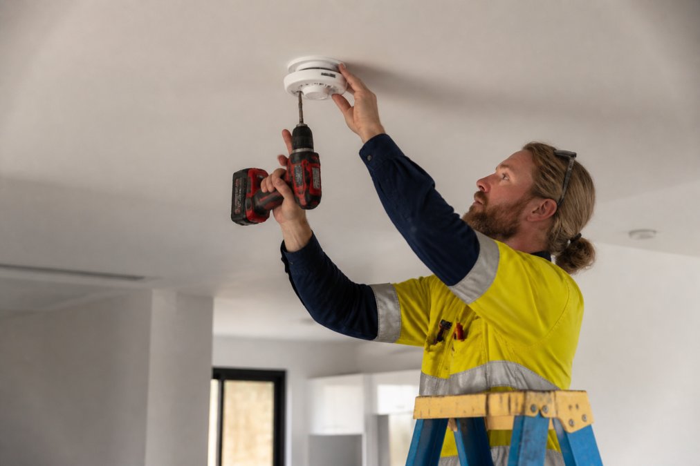 Qualified electrician installing a hardwired smoke alarm on a residential ceiling using a drill and safety equipment, wearing high-visibility clothing in a well-lit modern home