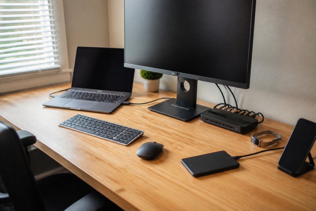 Organized remote work desk with multiple connected peripherals, docking station with visible cables neatly managed, laptop charging, external monitor, and wireless mouse positioned for ergonomic access